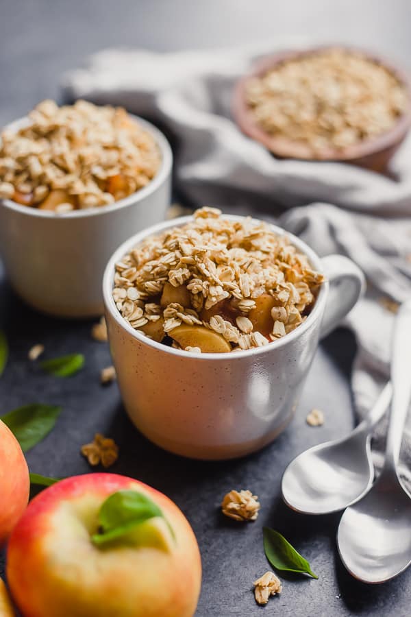 mugs of lazy girl apple crisp, some spoons, some granola, and some apples