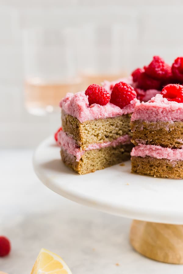 a slice of vegan lemon raspberry cake on a cake stand with two wine glasses behind it