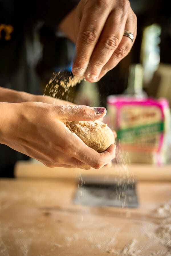 a hand holding a ball of pasta dough and another hand sprinkling flower on top