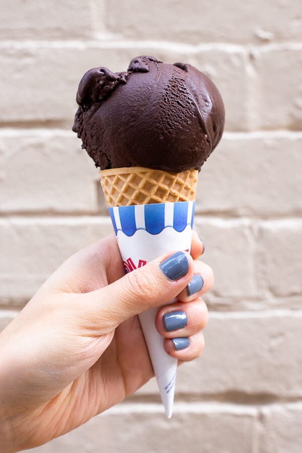 an ice cream cone with a scoop of Vegan Dark Chocolate Ice Cream in front of a white brick wall