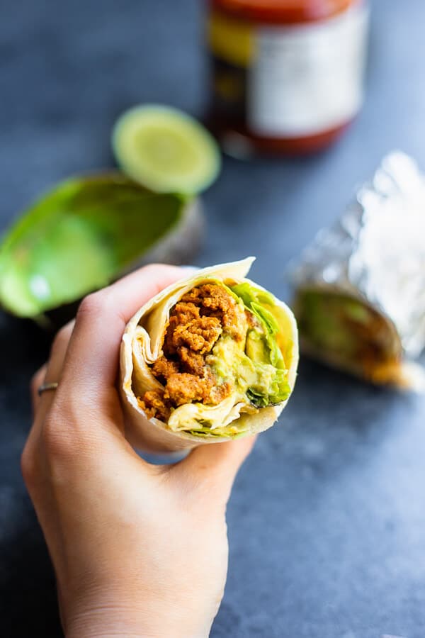 hand holding a vegan seitan burrito with ingredients in the background