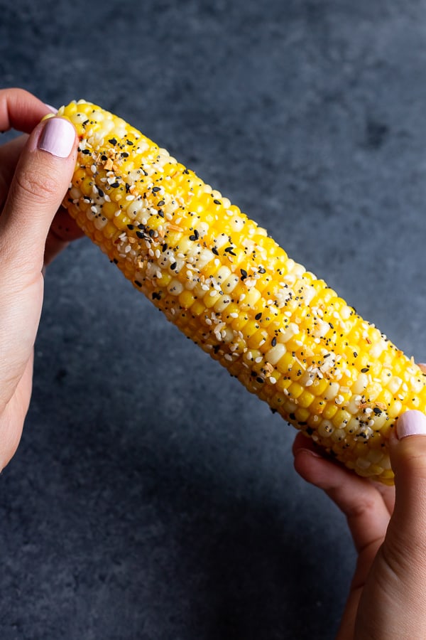two hands holding a corn on the cob that's covered in everything bagel seasoning