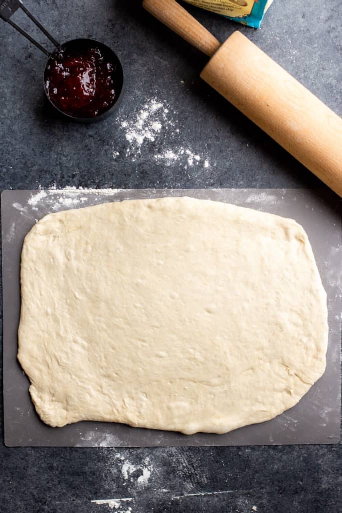 Vegan sweet roll dough rolled out on a cutting board with a rolling pin, a cup of strawberry preserves, and a bag of flour