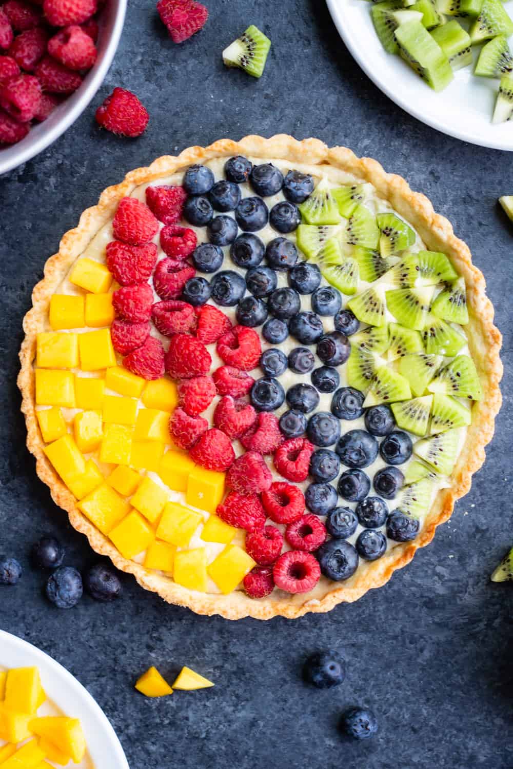 a vegan cream cheese fruit tart with mangos, raspberries, blueberries, and kiwi with bowls of the fruit surrounding it