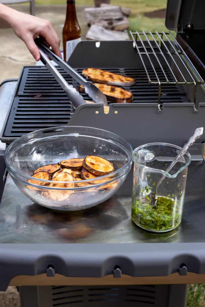 a bowl of grilled potatoes next to a jar of herb oil and grilled bread on the grill