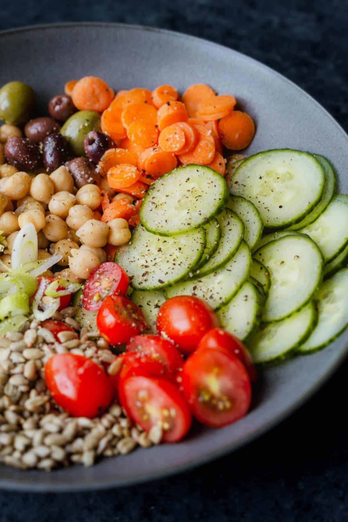 a close-up of a lettuceless salad of cucumbers, tomatoes, sunflower seeds, green onions, chickpeas, olives, and carrots in a dark grey bowl