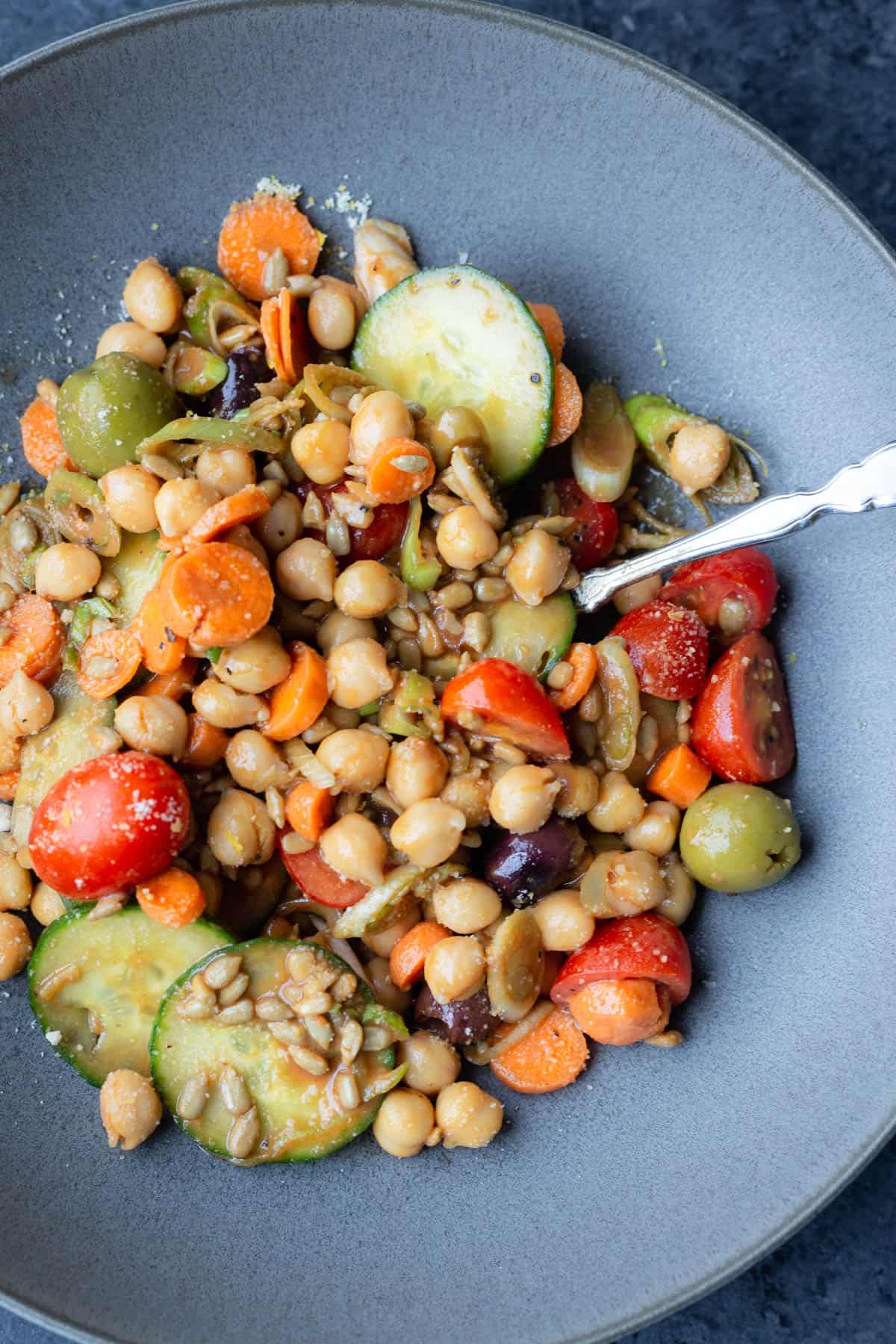 a mixed up bowl of a lettuceless salad of cucumbers, tomatoes, sunflower seeds, green onions, chickpeas, olives, and carrots